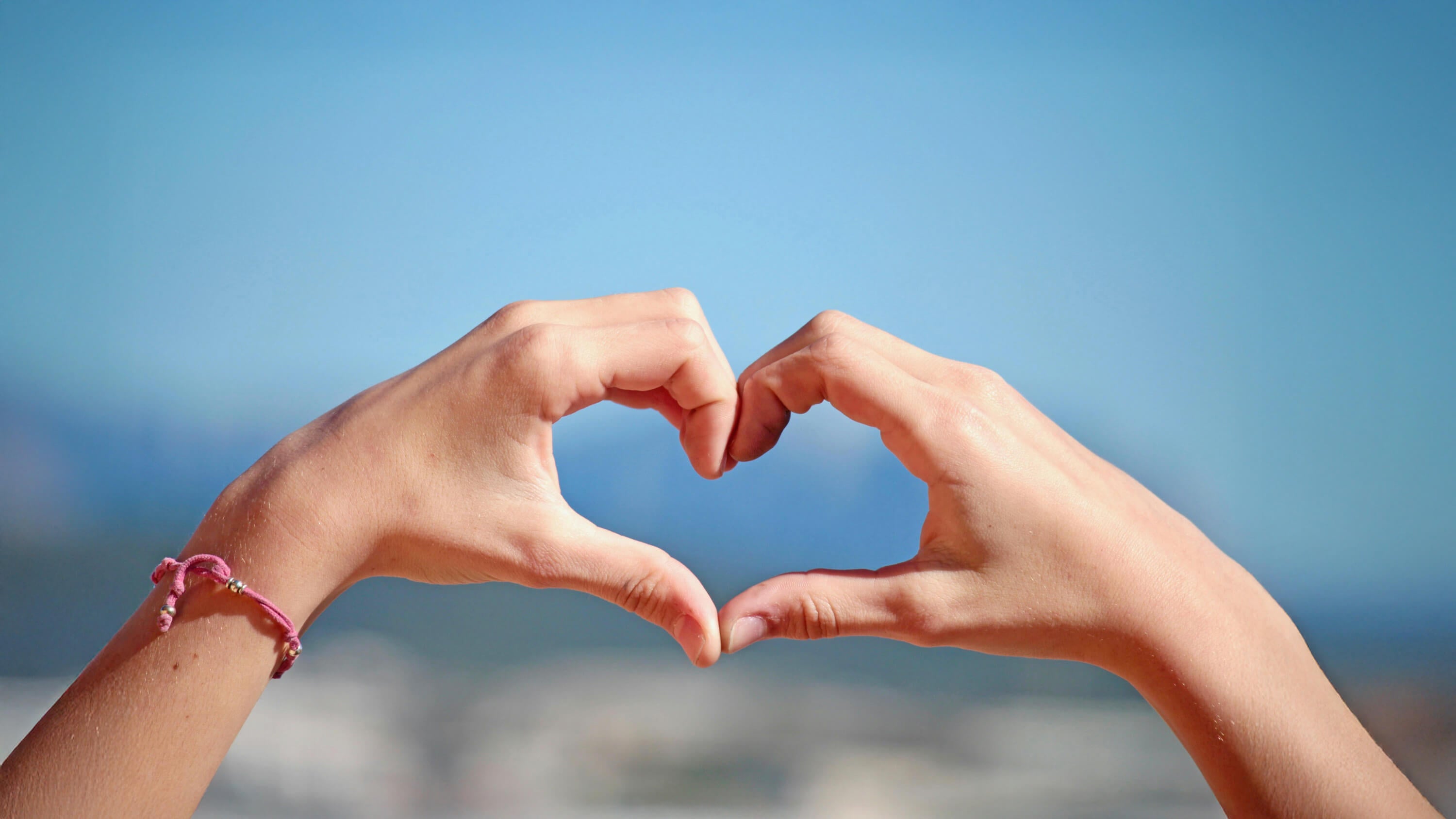 Two hands making a heart shape against a blurred beach background